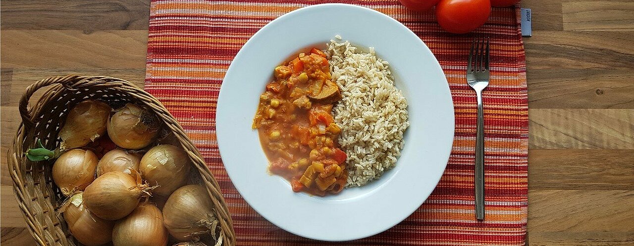 Auf dem Bild ist ein Teller mit Curry zu sehen. Unten links im Bild ist ein Korb mit Zwiebeln, oben rechts liegt ein Strauch Tomaten. 