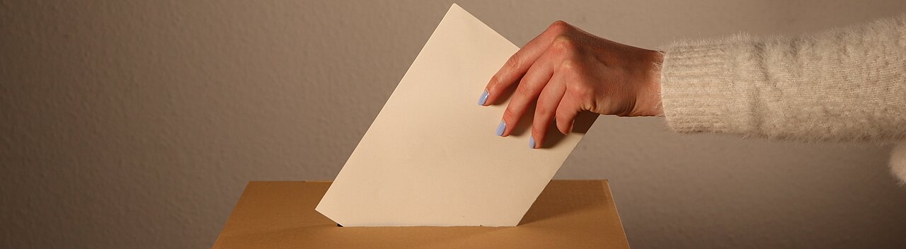 Man sieht eine Hand die einen gefalteten Zettel in eine Wahlurne steckt. 