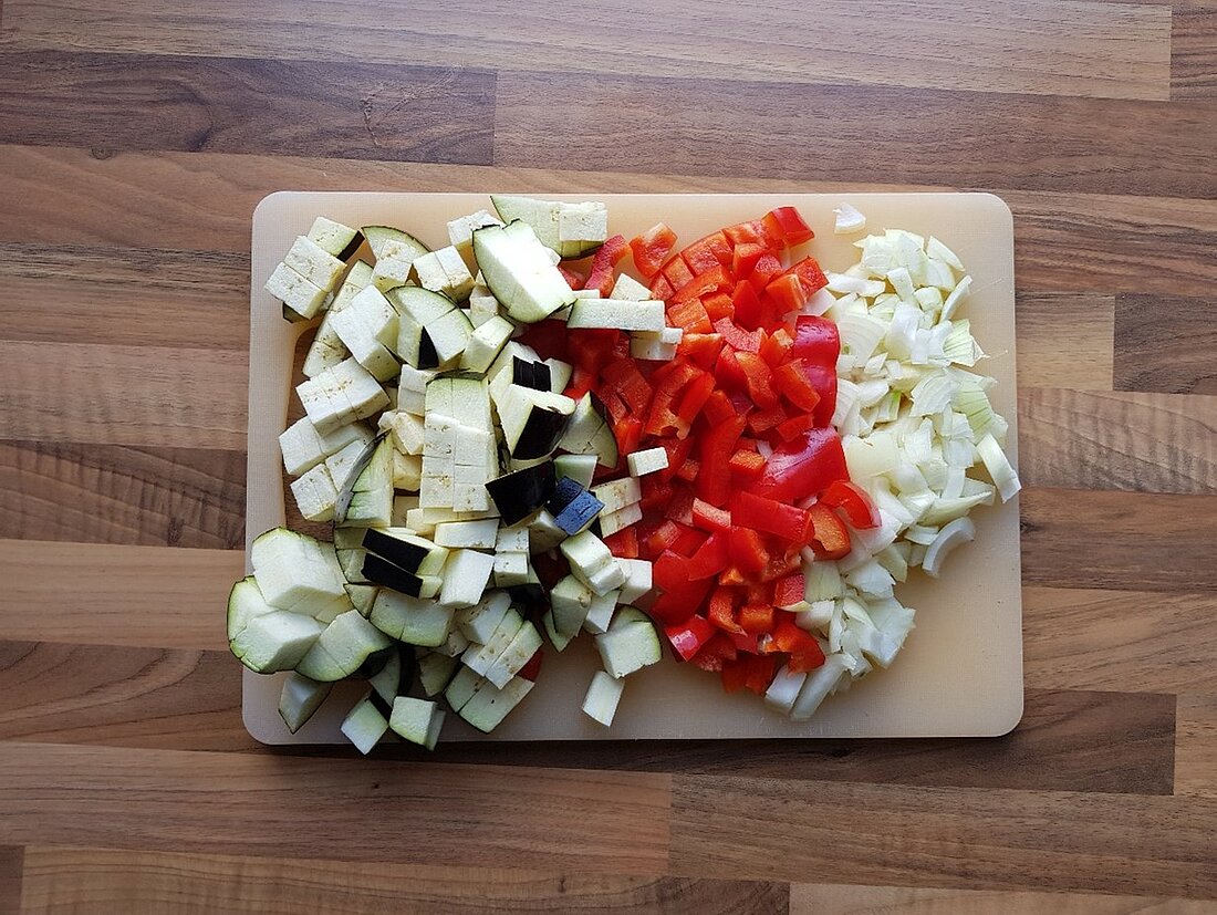 Auf dem Bild ist ein Schneidebrett zu sehen, auf dem Aubergine, Paprika und Zwiebeln liegen. 