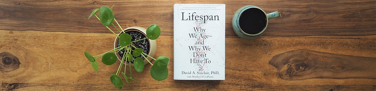 Das Buch "Lifespan: Why we Age - and Why We Don't Have To" liegt auf einem Holztisch. Daneben stehen eine Pflanze und eine Tasse. 