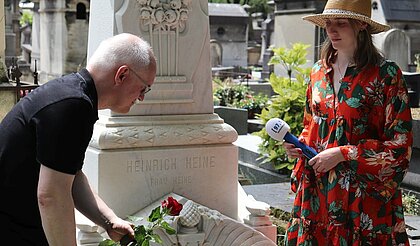 Wir sehen Heines Grab in Paris. Ein Redakteur des Hochschulradios legt eine Rose nieder. Eine Redakteurin steht mit dem Aufnahmegerät neben dem Grab.