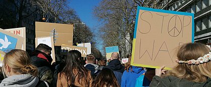 Zu sehen sind Demonstrant:innen, die gegen den Krieg auf der Straße sind. Zu sehen ist u.a. ein Plakat mit der Aufschrift "Stop War".