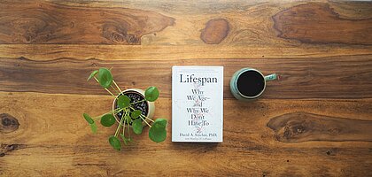 Das Buch "Lifespan: Why we Age - and Why We Don't Have To" liegt auf einem Holztisch. Daneben stehen eine Pflanze und eine Tasse. 