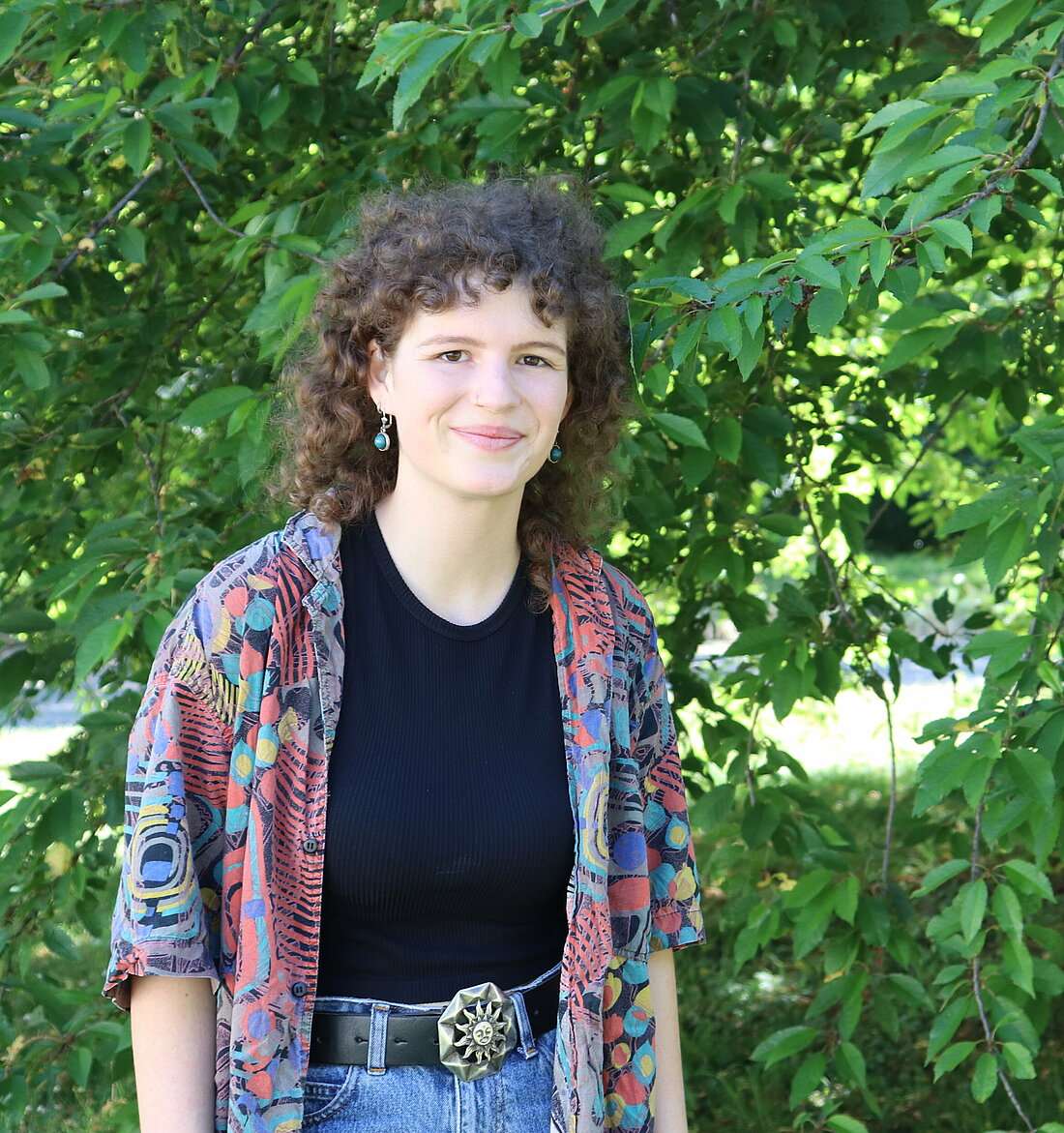 Die Spitzenkandidatin der Linken Liste studiert Sozialwissenschaften an der HHU (Foto: Anna Urban) Zu sehen ist eine junge Frau mit braunen Harren, die vor einer Grünfläche steht und lächelt