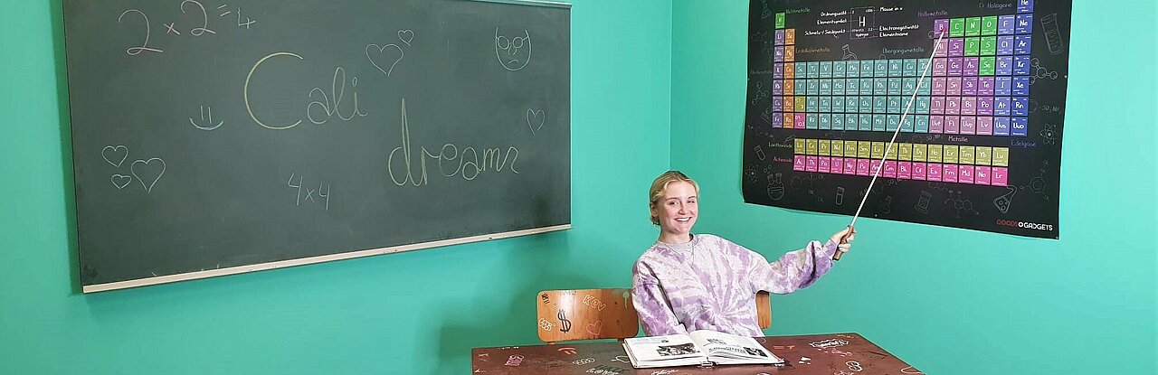 Auf dem Bild sieht man die Autorin im Klassenzimmer auf ein Plakat mit dem Periodensystem zeigen.