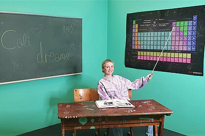 Auf dem Bild sieht man die Autorin im Klassenzimmer auf ein Plakat mit dem Periodensystem zeigen.