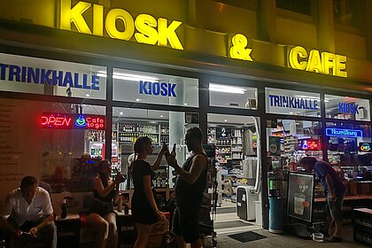 Man sieht einen Kiosk bei Nacht von aussen. Davor stehen zwei Menschen die sich zuprosten.