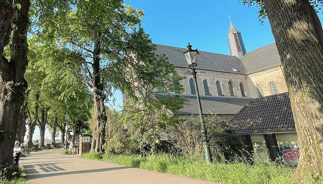 Es ist ein Ausschnitt der Kaiserswerther Rheinuferpromenade vor der St. Suitbertus Basilika zu sehen.