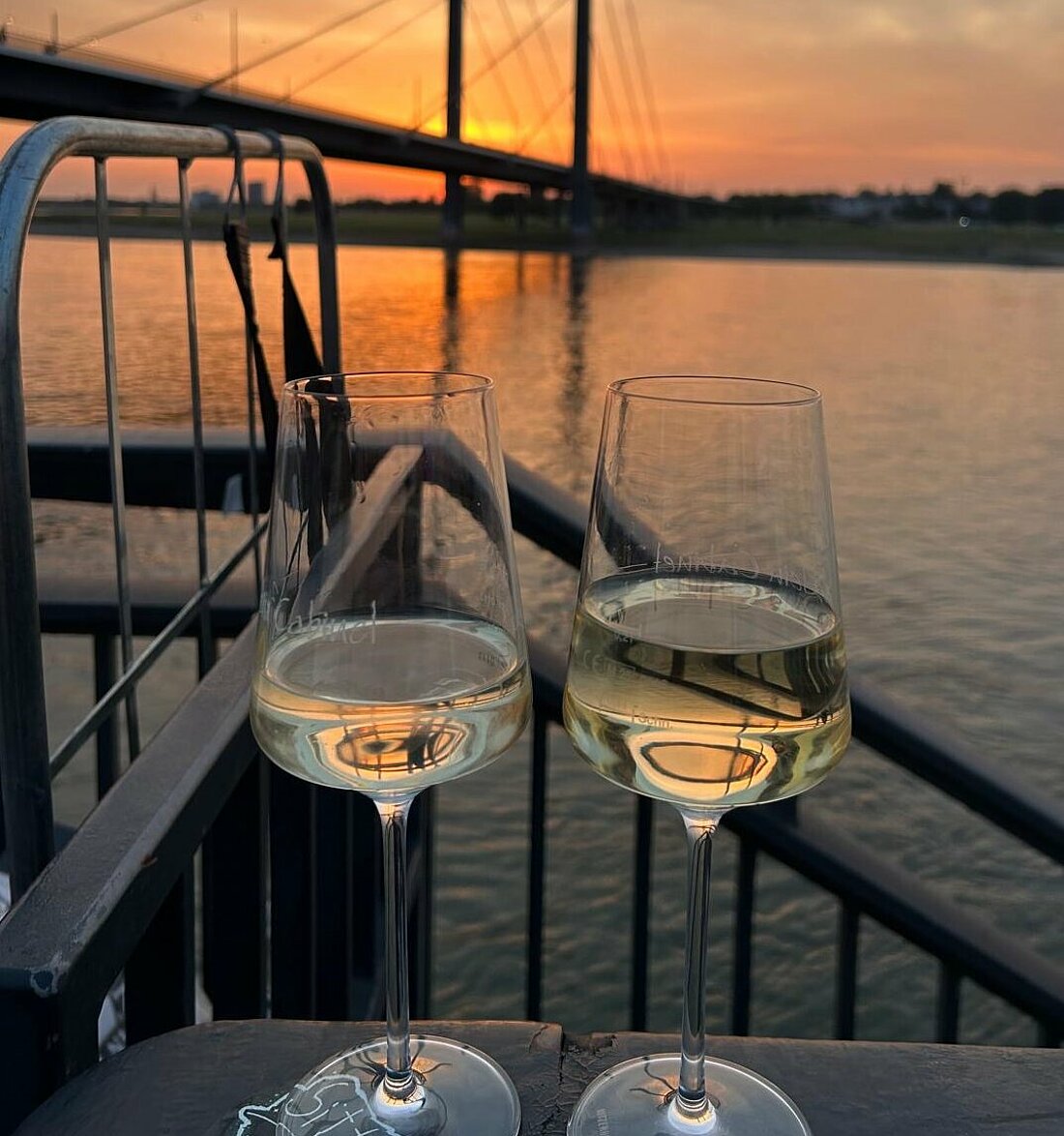 Auf dem Bild sieht man zwei Gläser mit Wein, die vor dem Rhein stehen bei einem Sonnenuntergang.