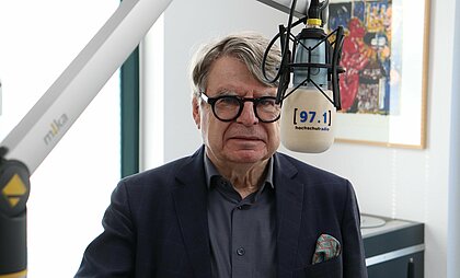 Auf dem Foto ist Ulrich von Alemann zu sehen. Er steht neben dem Mikrofon und lächelt in die Kamera