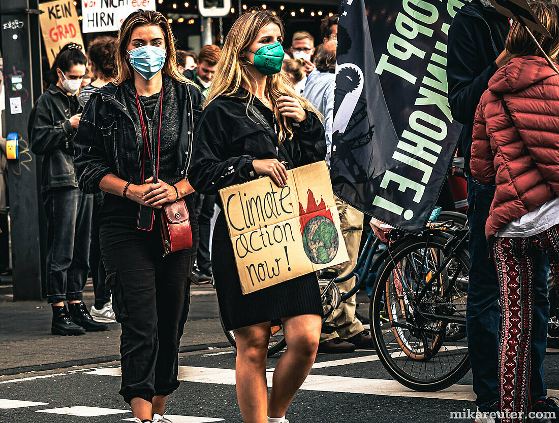 Auf dem Bild sieht man eine Fridays for Future Demonstration in Düsseldorf. Im Mittelpunkt des Bildes sind zwei junge Frauen, die eine medizinische Maske tragen und ein Schild in den Händen halten auf dem steht: "climate action now". Im Hintergrund sind noch weitere Demoteilnehmer:innen zu sehen.