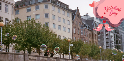 An der Düsseldorfer Rheinpromenade schweben Seifenblasen vor Bäumen und Wohnhäusern. Oben im Bild ist ein „Valentine’s Day“-Schriftzug, darunter zwei küssende Vögel und zwei Herzen.