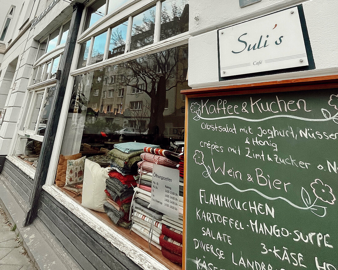  Es ist die Hauswand des Cafe Sulis mit Fokus auf das dort angebrachte Namensschild und der Menü-Tafel zu sehen.