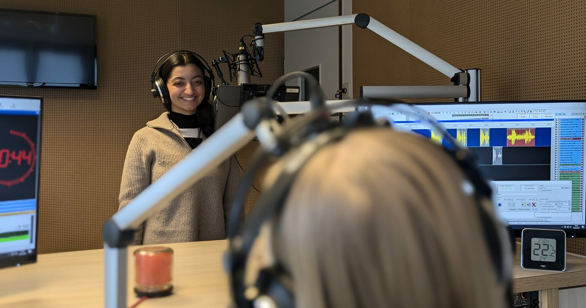 Auf dem Foto ist Sita Grabbe vom AStA-Vorstand der RSH zu sehen. Sie steht am Mikrofon und lächelt in die Kamera. Im Vordergrund sieht man verschwommen den Hinterkopf von der Moderatorin. Vor ihr sind zwei Bildschirme zu sehen.