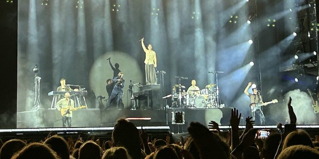 Man sieht die Bandmitglieder auf der Bühne. Der Sänger steht auf dem Klavier.