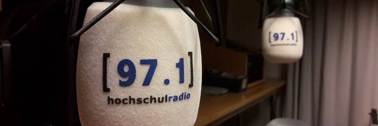 In einem Radiostudio hängen sich zwei Mikrofone gegenüber. Auf beiden steht 97.1 hochschulradio.