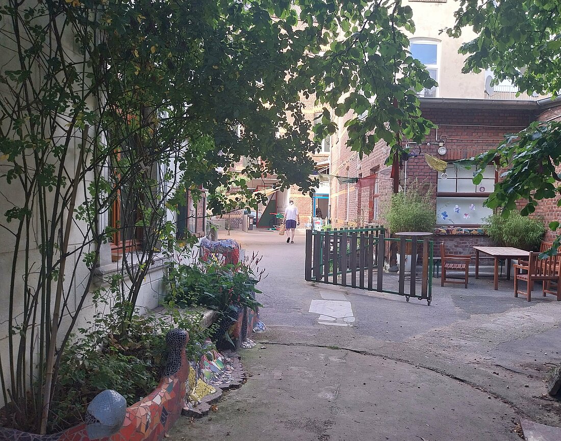 Ein Innenhof, der von grünen Bäumen eingerahmt ist. Links steht ein Tisch mit Stühlen, im Hintergrund geht ein Mann in Richtung Straße. 
