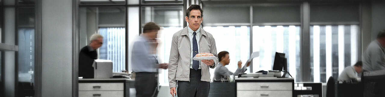 Zu sehen ist Walter Mitty, die Hauptfigur des Films, in einer typischen Büroszene. Er steht ruhig in der Mitte des Raums, während um ihn herum andere Personen in Bewegung sind.