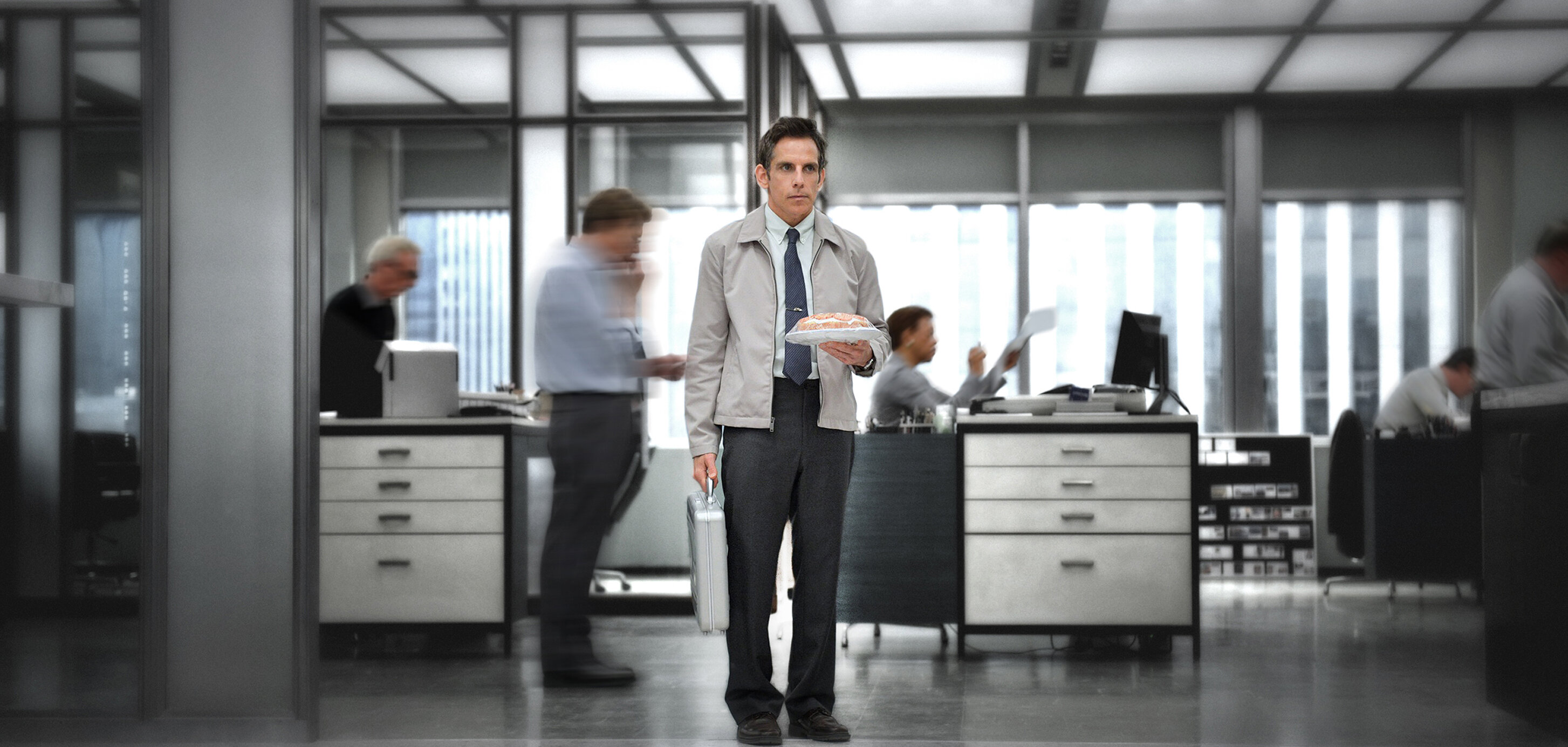 Zu sehen ist Walter Mitty, die Hauptfigur des Films, in einer typischen Büroszene. Er steht ruhig in der Mitte des Raums, während um ihn herum andere Personen in Bewegung sind.