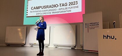 Man sieht die Bühne beim Campusradiotag auf der die Moderatorin das Publikum begrüßt