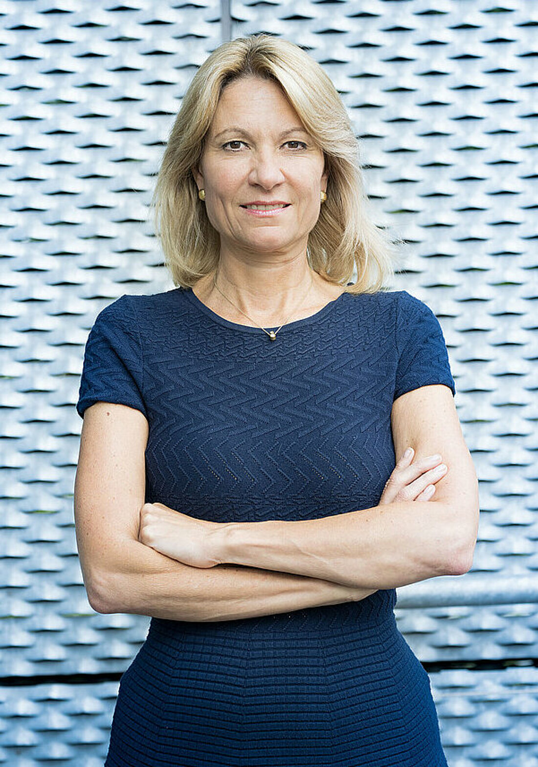 Auf dem Bild ist Prof. Anja Steinbeck, die Rektorin der Heinrich-Heine-Universität zu sehen. Sie steht mit überkreuzten Armen vor einem grauen Gebäude.