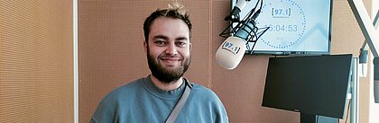 Auf dem Foto ist Elias Warnke zu sehen. Er steht neben einem Mikrofon und lächelt in die Kamera.