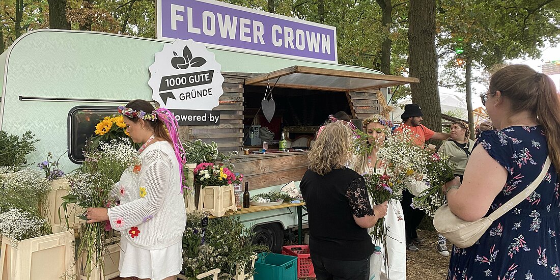 Auf dem Foto ist ein grüner Wohnwagen mit der Aufschrift "Flower Crown" zu sehen. Davor stehen viele Blumenstäuße in Kästen und Menschen.