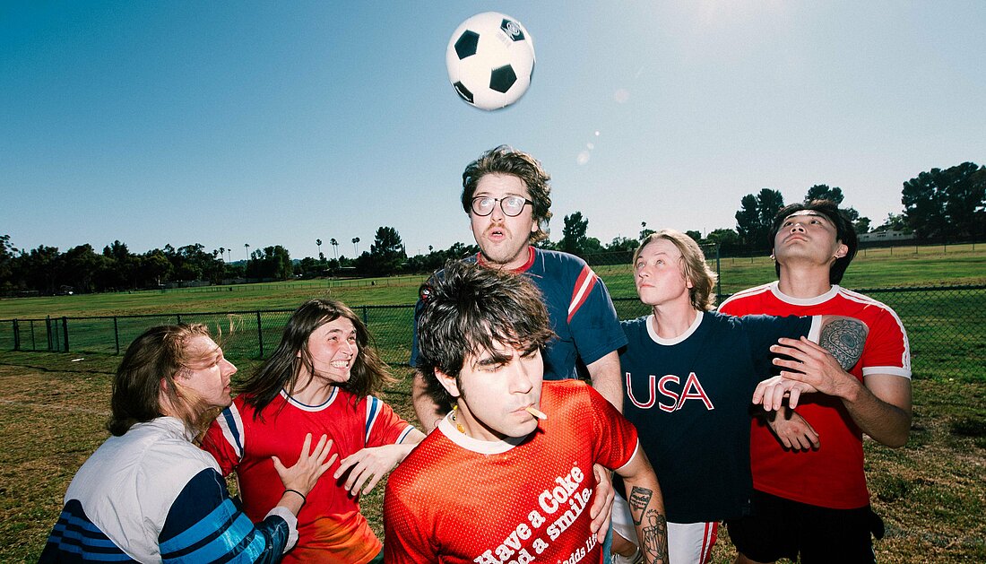 Auf dem Foto sieht man die Band The Sophs. Sie stehen auf einem Feld und spielen Fußball, der Fußball fliegt in der Luft
