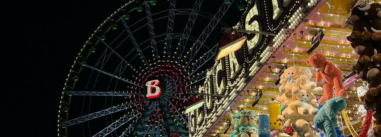 Auf dem Foto sieht man die Kirmes bei Nacht. Im Hintergrund ist das Riesenrad beleuchtet und vorne ist ein Stand ebenfalls beleuchtet