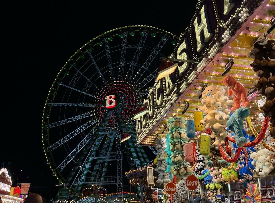 Auf dem Foto sieht man die Kirmes bei Nacht. Im Hintergrund ist das Riesenrad beleuchtet und vorne ist ein Stand ebenfalls beleuchtet