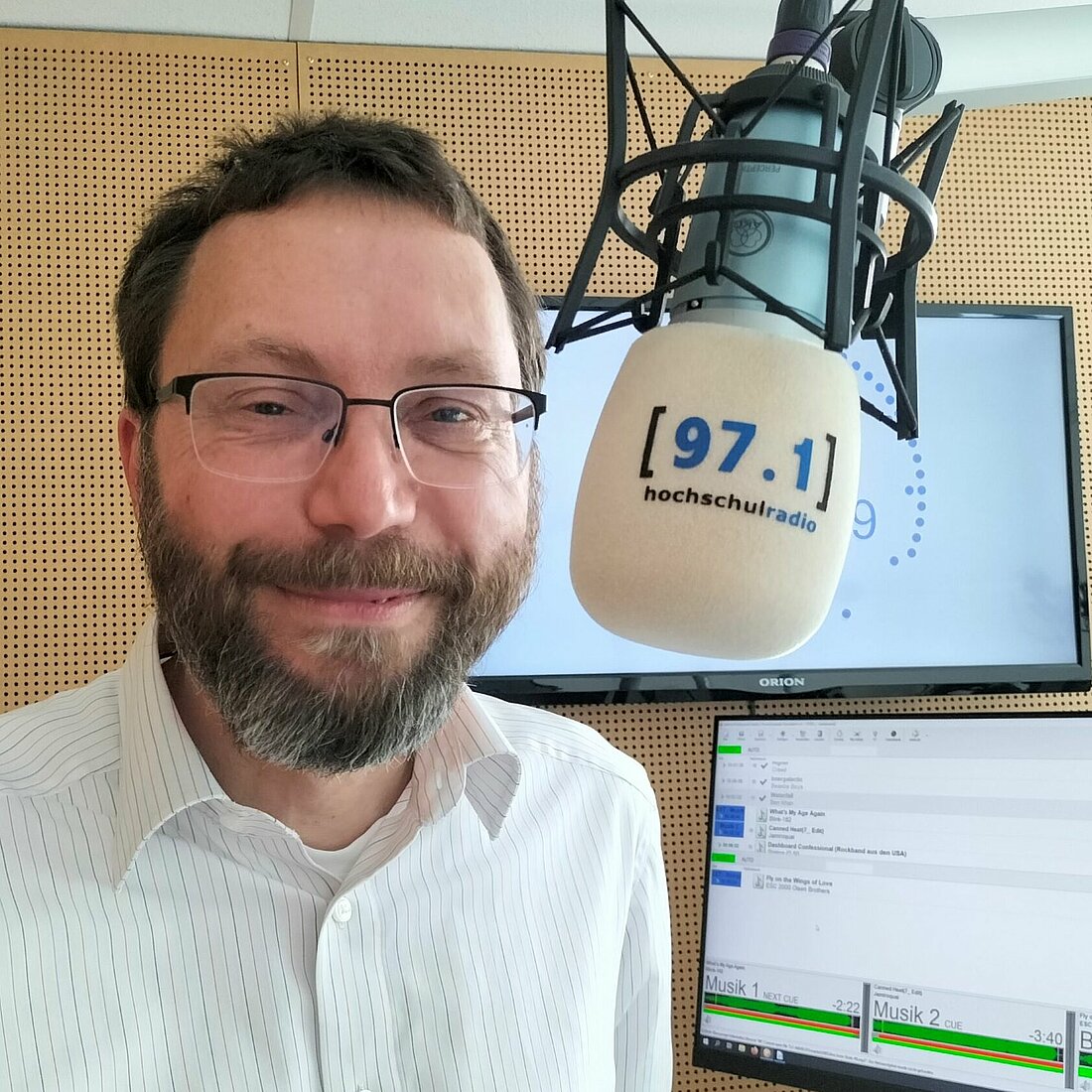 Auf dem Foto ist Stefan Adler zu sehen. Er steht vor einem Mikrofon und lächelt in die Kamera. Hinter ihm sind Teile des Studios, wie ein Computerbildschirm und eine Uhr zu erkennen.