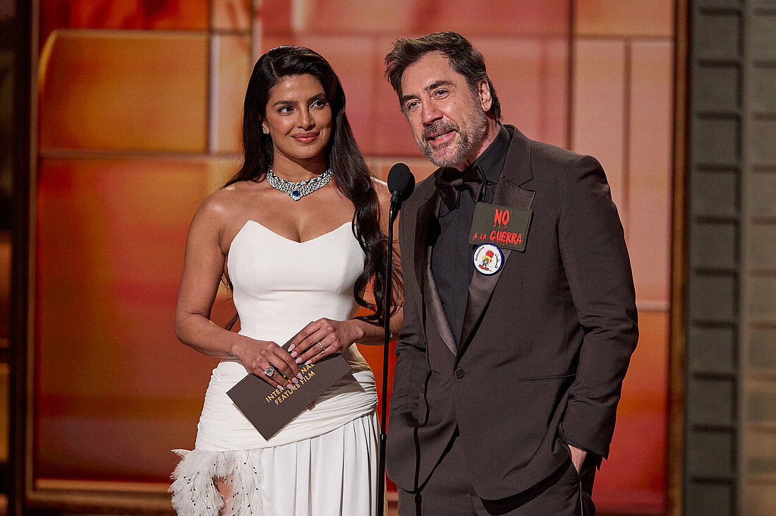 Priyanka Chopra Jonas und Javier Bardem stehen gemeinsam auf der Bühne des Dolby Theater. Bardem trägt einen Anstecker gegen Krieg an seinem Jackett.