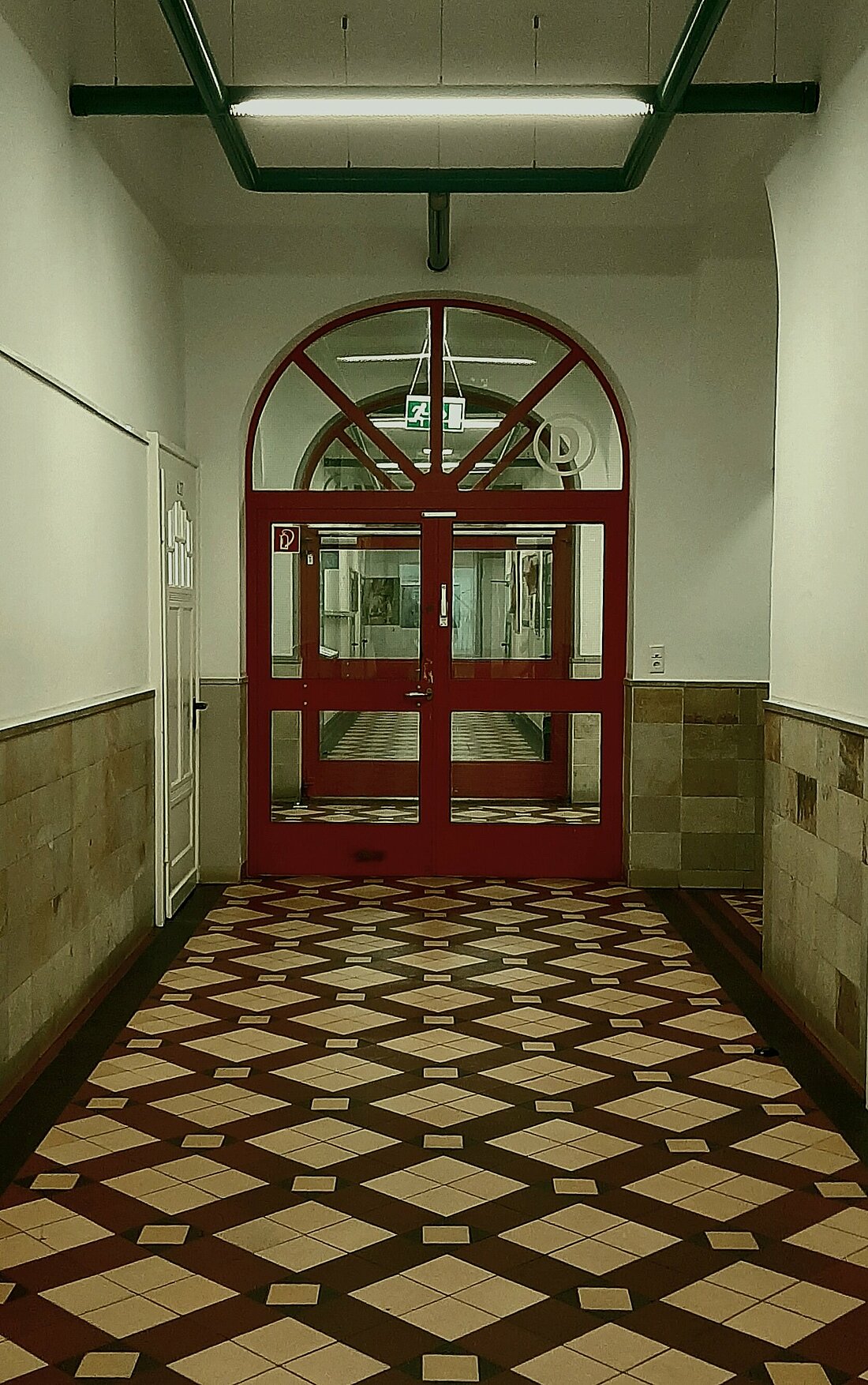 Auf dem Bild sieht man einen schmalen Gang mit kariertem Fliesenboden und gelblichen Wänden. An seinem Ende befindet sich eine rote Glastür mit einer Leuchtstofflampe darüber.