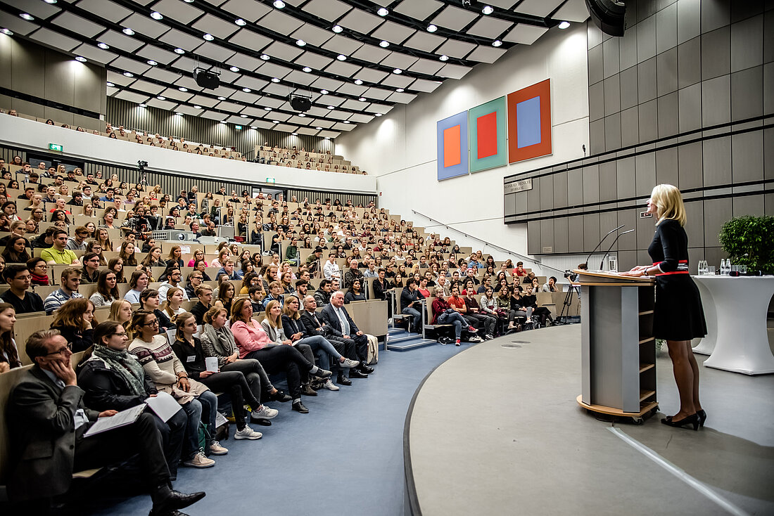 Prof. Dr. Anja Steinbeck spricht vor einem Hörsaal voller Erstsemester:innen.