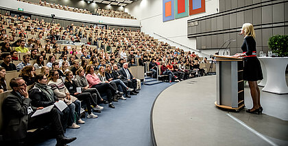 Prof. Dr. Anja Steinbeck spricht vor einem Hörsaal voller Erstsemester:innen.