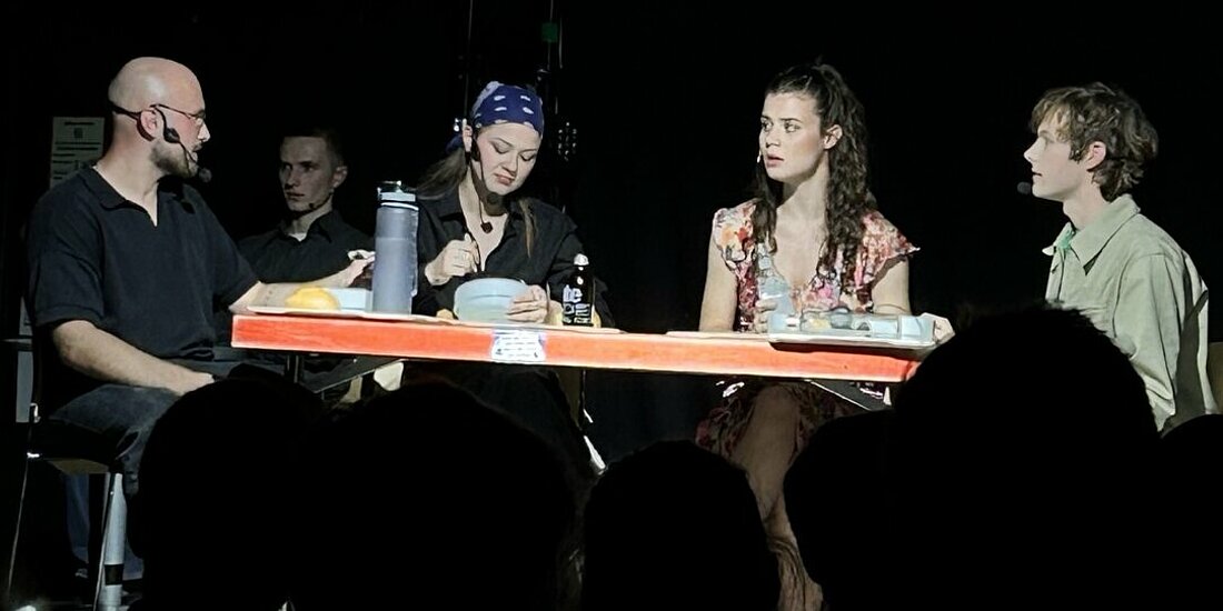 Man sieht die drei Hauptfiguren des Mensa-Musicals, wie sie an einem Tisch sitzen, essen und reden.