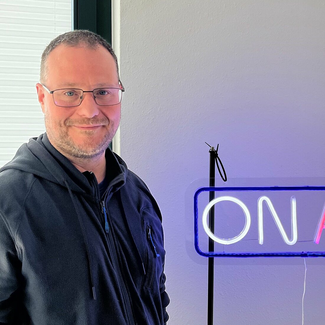 Auf dem Foto steht Boris S. Schmidt neben einem leuchtenden OnAir Schild und lächelt in die Kamera.