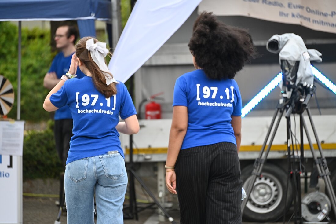 Auf dem Bild sieht man zwei Mädchen von hinten, die auf den Stand des Hochschulradios zugehen.