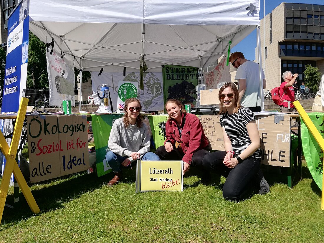 Auf dem Foto sieht man einen Infostand von den Students for future. Im Vordergrund sieht man drei Aktivistinnen von Students for future, die vor dem Infostand hocken mit einem Schild in der Hand auf dem steht „Lützerath bleibt!“ 