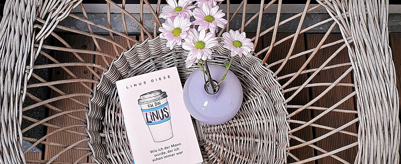 Zu sehen ist das Buch "Ich bin Linus" auf einem Korbstuhl. Rechts vom Buch befindet sich eine lila Blumenvase mit rosa Blumen.