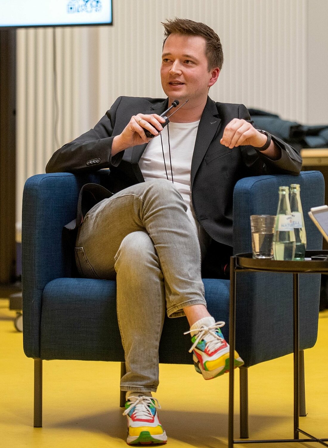 Auf dem Foto ist Daniel Hagemann auf einer Podiumsdiskussion zu sehen. Er sitzt auf einem blauen Sessel und spricht in ein Mikrofon. 