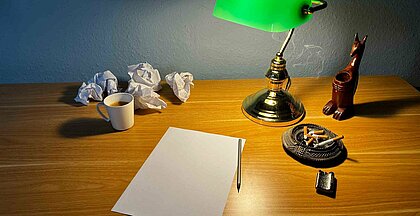 Auf dem Bild sieht man einen Schreibtisch mit einem Papier, einer Lampe und Zigaretten drauf. In der Ecke steht noch eine Tasse Kaffee (Foto: Gregory Sheftel) 