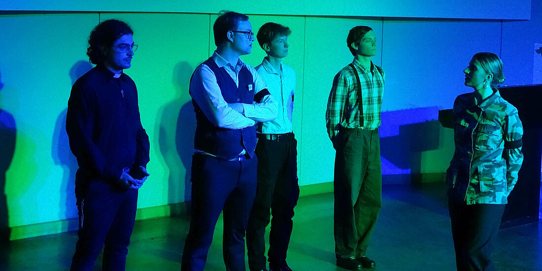 Auf dem Bild sind vier Schauspieler zu sehen, die aufgestellt in einer Reihe stehen. Ihnen zugewandt steht eine weitere Schauspielerin.