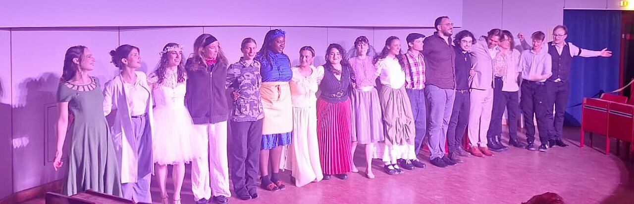 Auf dem Bild sind alle Schauspieler:innen des Ensembles zu sehen. Sie stehen alle aufgereiht auf der Bühne, um sich zu verbeugen. 