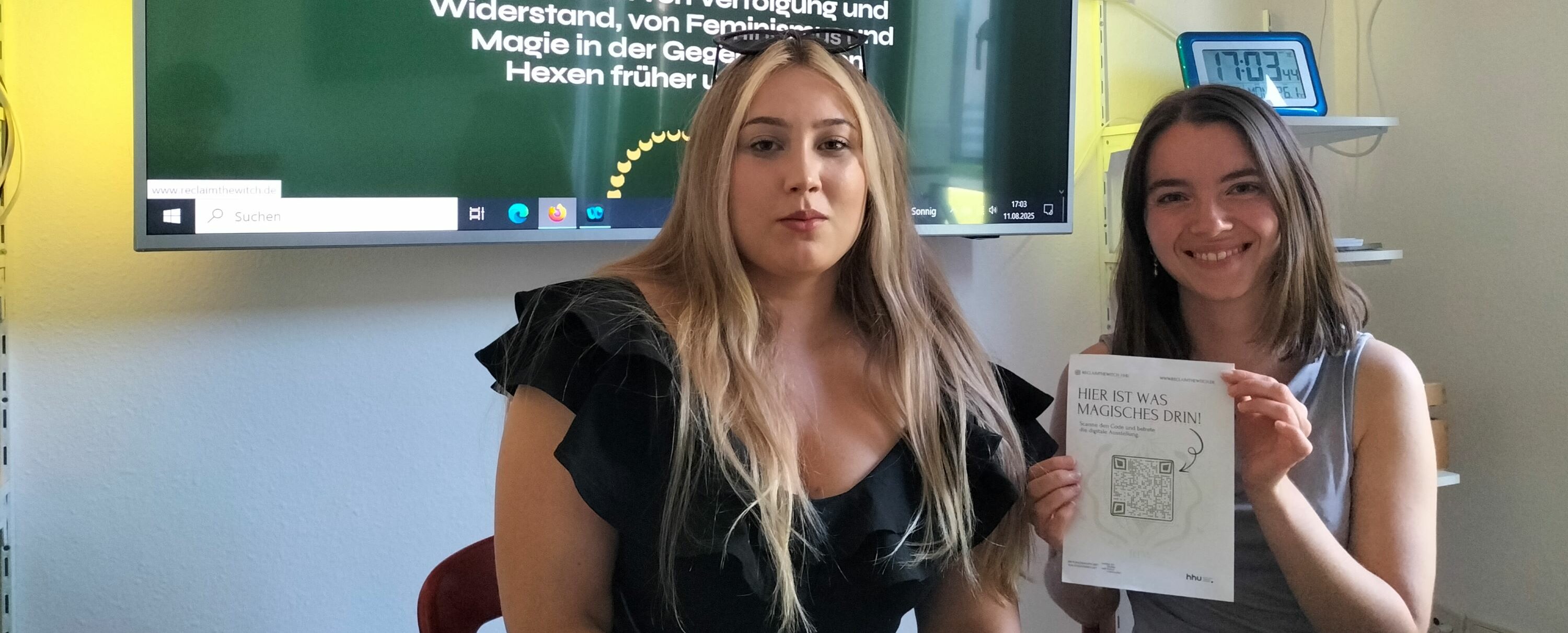 Auf dem Foto sind Nora und Blanca zu sehen. Sie sitzen nebeneinander und lächeln in die Kamera. Nora hält ein Flyer zu der Ausstellung hoch.