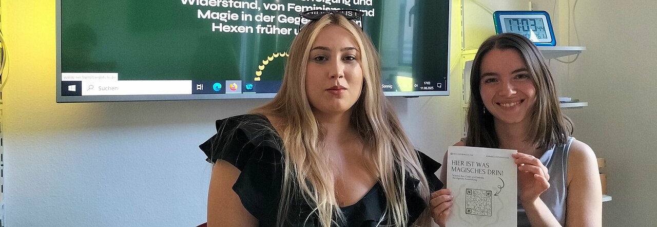 Auf dem Foto sind Nora und Blanca zu sehen. Sie sitzen nebeneinander und lächeln in die Kamera. Nora hält ein Flyer zu der Ausstellung hoch.