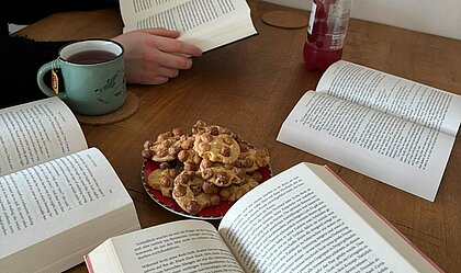 Eine Person sitzt an einem Tisch und liest. Auf dem Tisch liegen mehrere aufgeschlagene Bücher, ein Teller mit Keksen, eine Tasse Tee und eine Flasche Limonade.