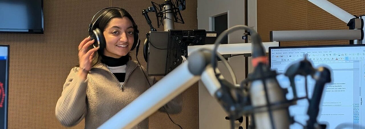 Auf dem Foto ist Sita Grabbe zu sehen. Sie steht im Studio vorm Mikrofon und lächelt in die Kamera.