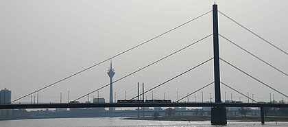 Auf dem Bild sieht man die  Rheinkniebrücke und den Fernsehturm in Düsseldorf. Über die Brücke fährt eine Ubahn.