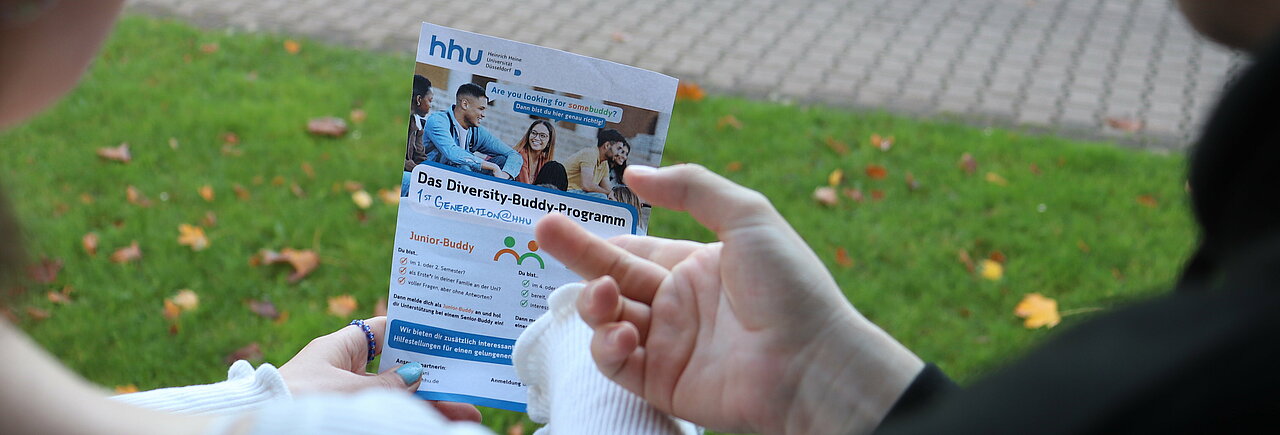 Auf dem Foto ist der Flyer vom Buddy-Programm zu sehen. Zwei Hände zeigen auf dem Flyer